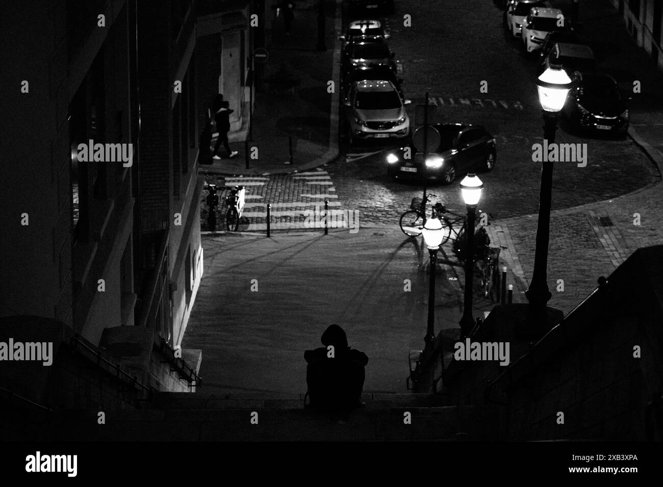 Schwarz-Weiß-Ansicht hinunter zu einer vermummten Figur, die auf Stufen neben einer Straßenlaterne auf einer gepflasterten Pariser Straße sitzt, nachts, 16. Arrondissement, Frankreich Stockfoto