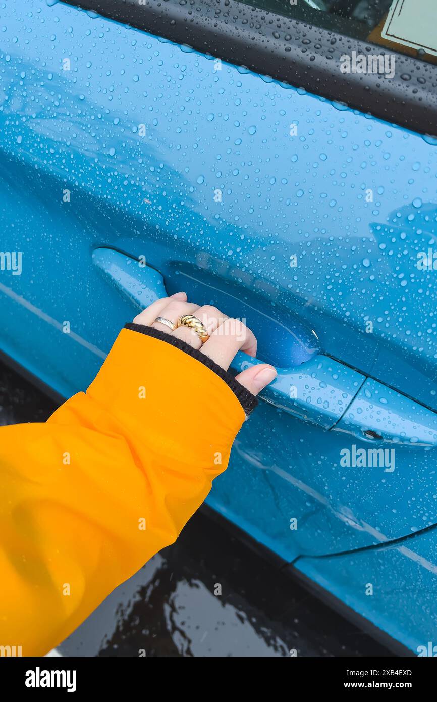 Weibliche Hand berührt den Griff eines blauen Autos in Nahaufnahme, Tür mit den Regentropfen Stockfoto