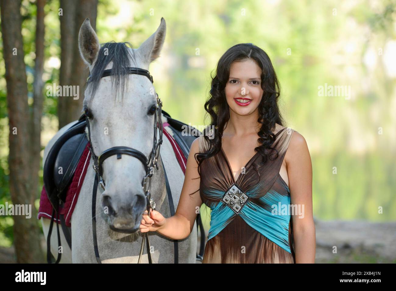 Eine Frau in braunem und blauem Kleid lächelt, während sie ein Quarab-Pferd in einem bewaldeten Gebiet in Bayern hält Stockfoto