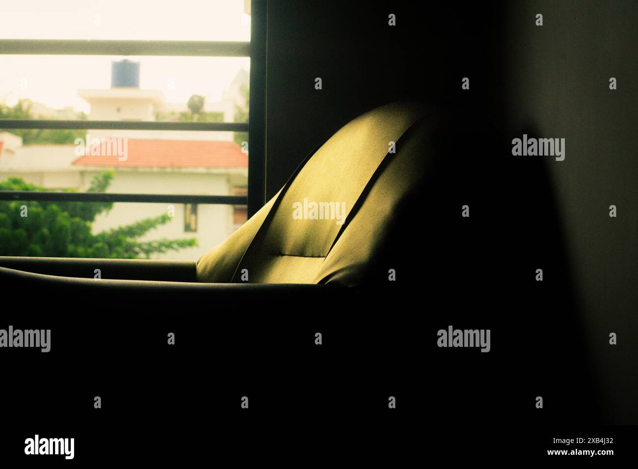 Ein einfacher Holzstuhl steht neben einem Fenster in einem schwach beleuchteten Raum. Die Fensterscheibe wirft ein schwaches Licht auf die Wand. Stockfoto