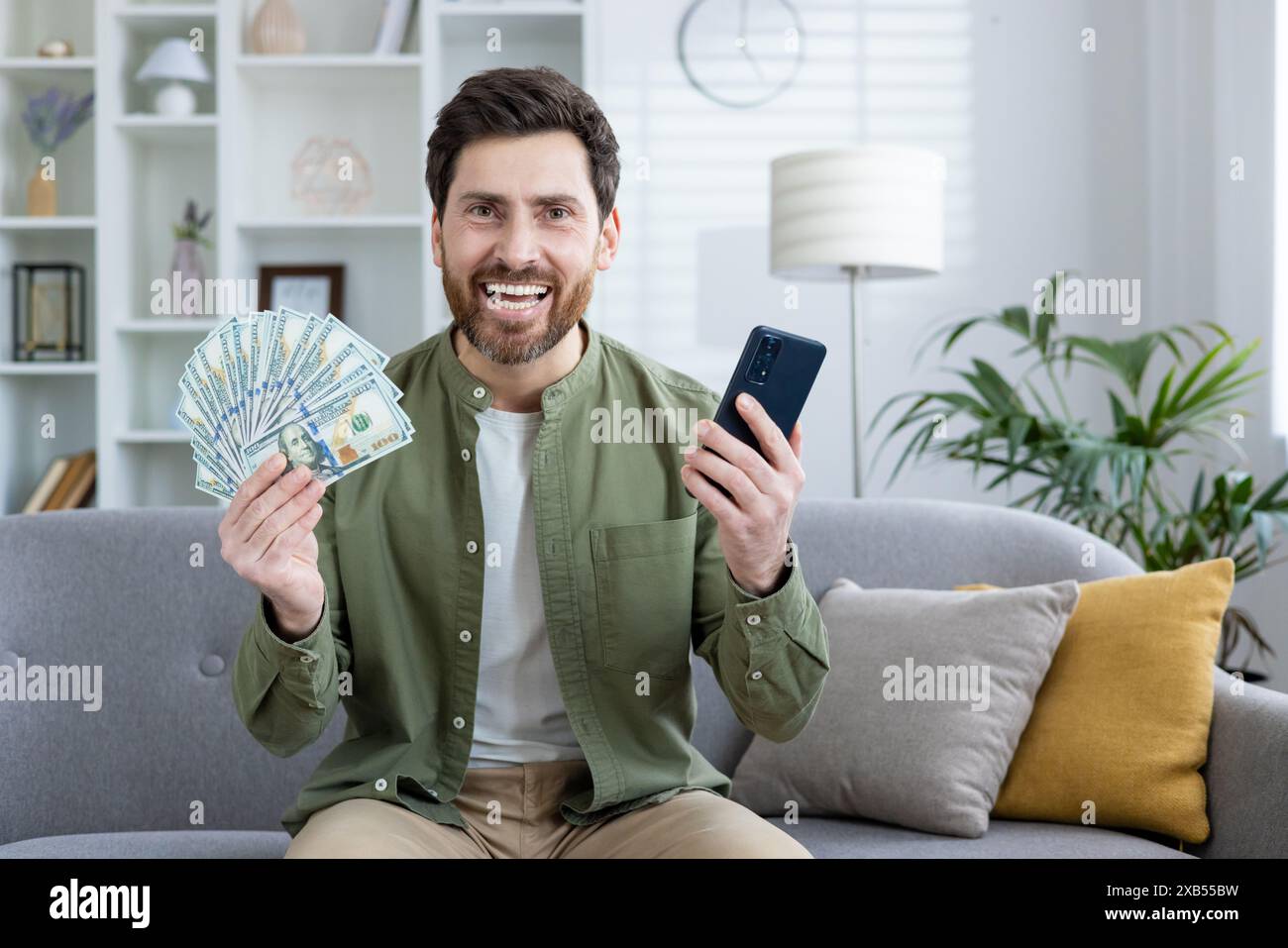 Porträt eines glücklichen jungen Mannes, der zu Hause auf dem Sofa sitzt und finanziellen Erfolg vor der Kamera genießt, Handy und Bargeld hält. Stockfoto