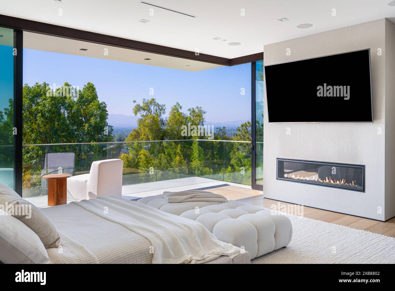 Ein modernes Schlafzimmer mit weißen Möbeln, großen Fenstern, Fernseher, Balkon mit Aussicht und weißer Bettwäsche Stockfoto