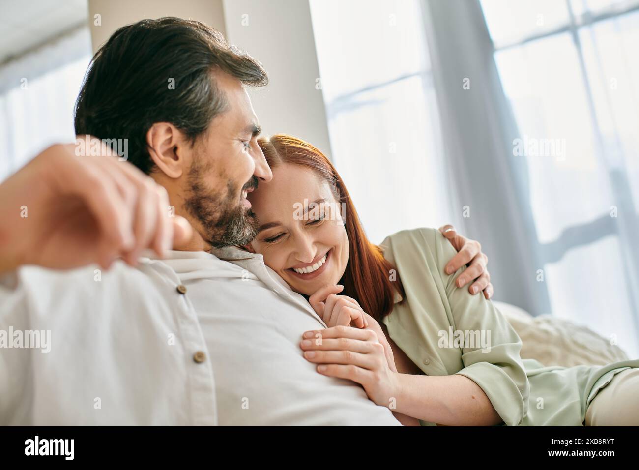 Eine rothaarige Frau und ein bärtiger Mann teilen sich eine warme Umarmung, während sie auf einer bequemen Couch in ihrer modernen Wohnung sitzen. Stockfoto
