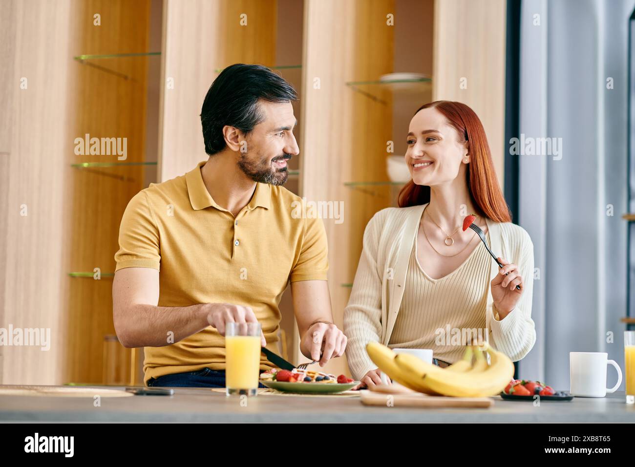 Ein bärtiger Mann und eine rothaarige Frau genießen ein gemütliches Frühstück in einer modernen Küche, was eine warme und harmonische Szene schafft. Stockfoto