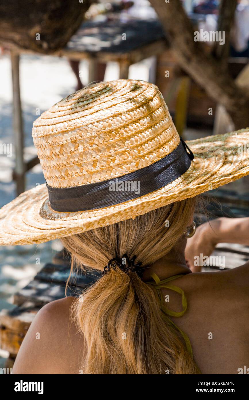 Nahaufnahme einer Frau mit langen blonden Haaren, die einen Strohhut mit schwarzem Band trägt und draußen in einem schattigen Bereich sitzt. Perfekt für Sommer, Entspannung, Stockfoto