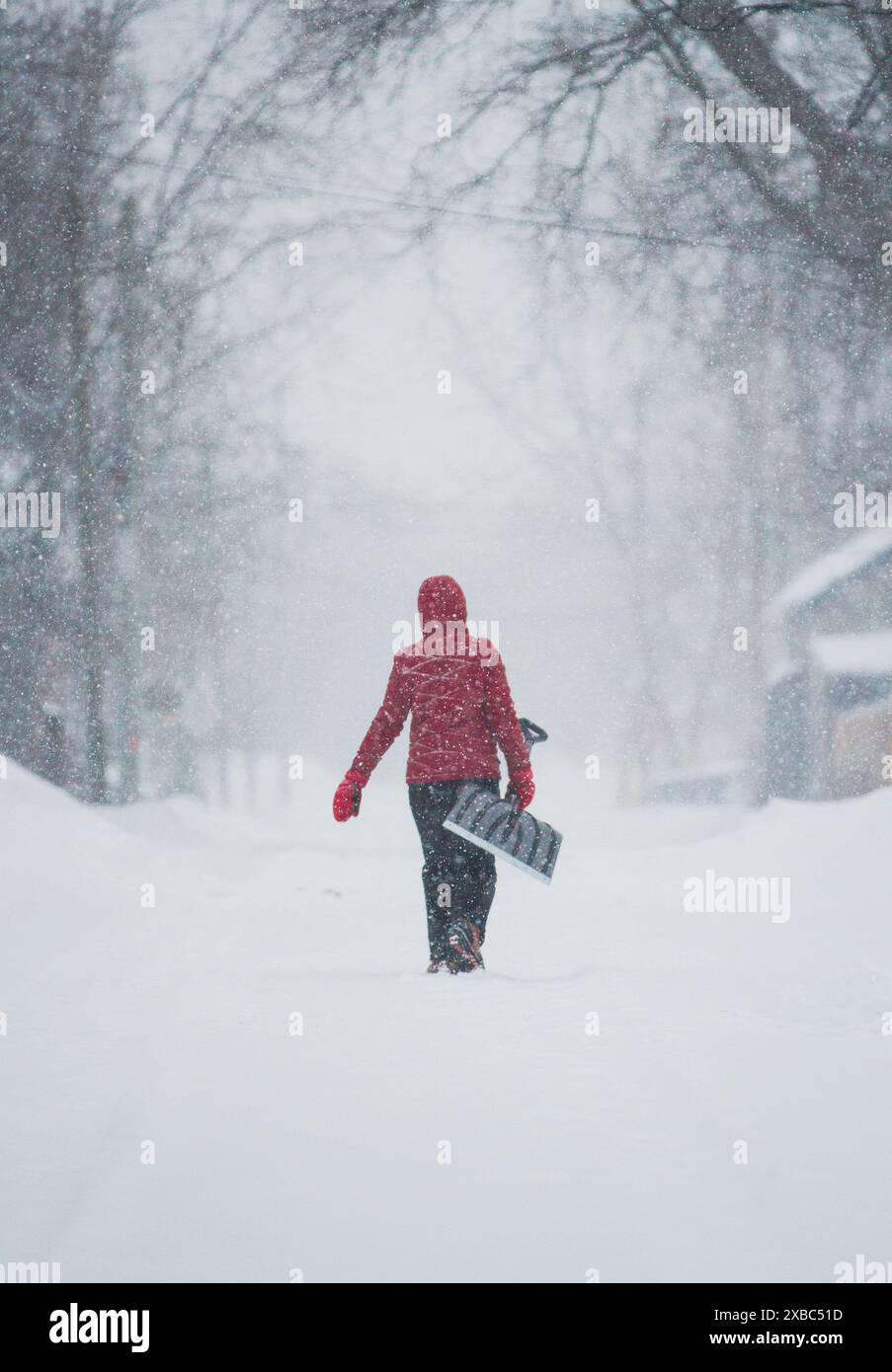 Eine Frau in rotem Mantel trägt eine Schneeschaufel auf einer Kleinstadtstraße mitten in einem Schneesturm Stockfoto