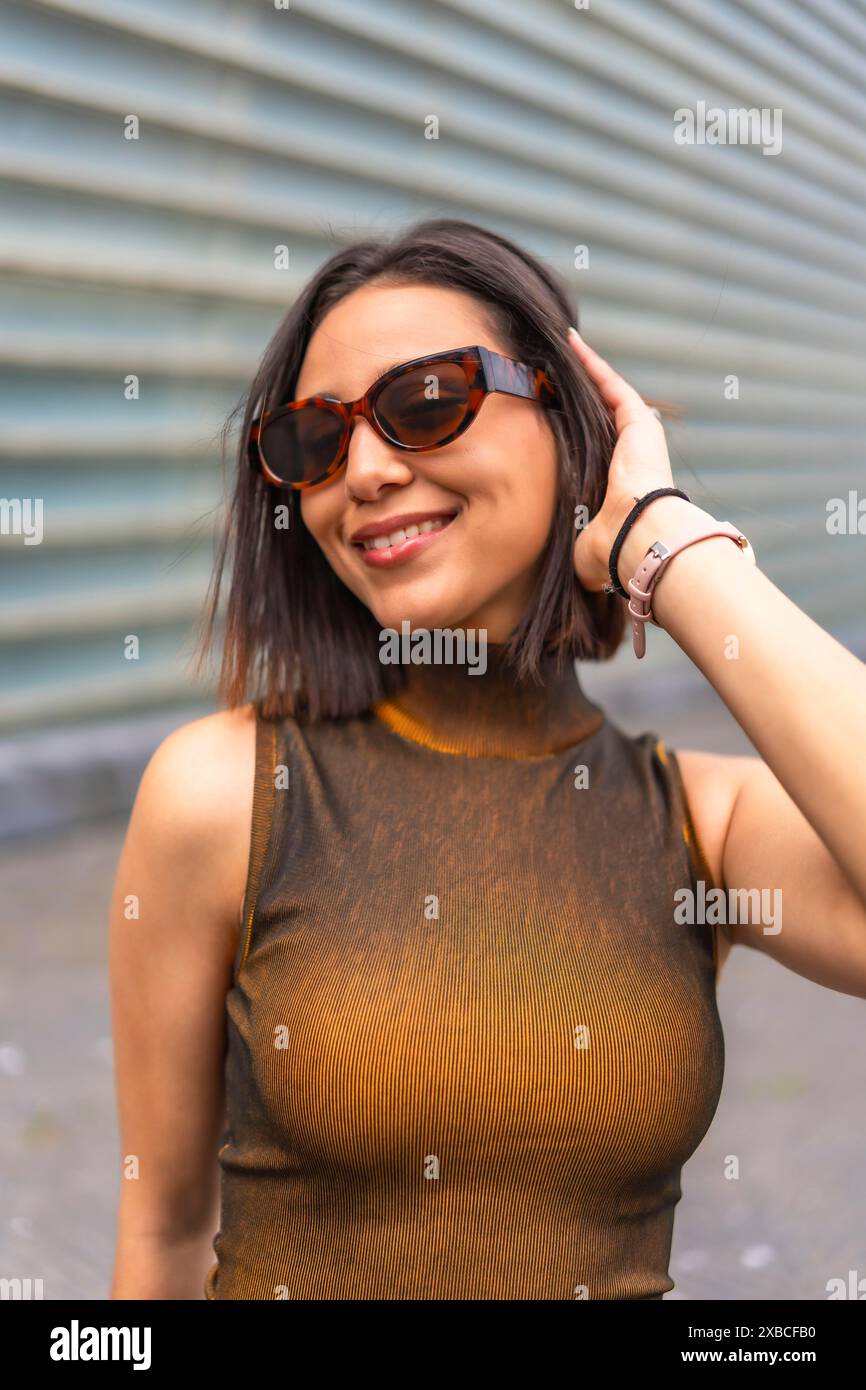 Eine Frau mit Sonnenbrille und braunem Oberteil mit einem Muster drauf. Sie lächelt und posiert für die Kamera Stockfoto
