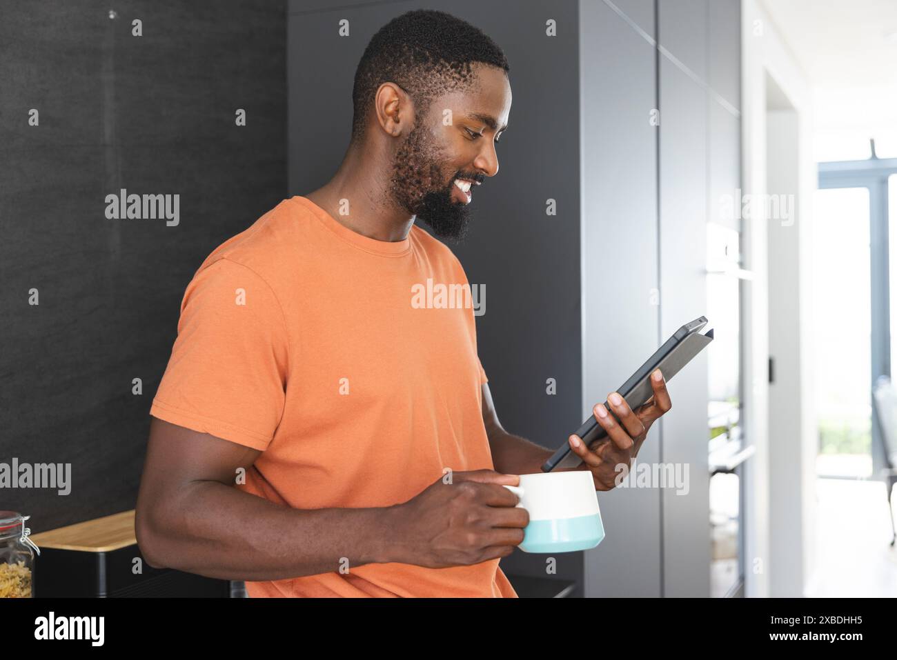Junger afroamerikanischer Mann mit Tablette und Kaffeetasse, lächelnd auf den Bildschirm. Er steht in einer modernen Küche mit Sonnenlicht, das durch die Large strömt Stockfoto
