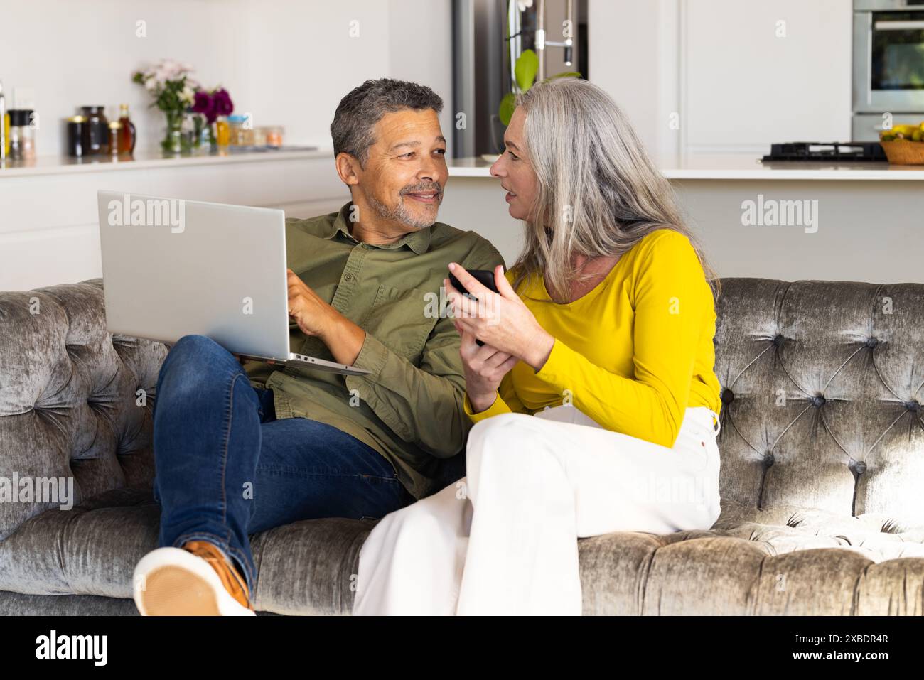Ein geschlechtsreiches Ehepaar sitzt zu Hause auf einer Couch, benutzt einen Laptop und ein Smartphone. Sie befinden sich in einem modernen, hellen Wohnzimmer mit einer Küche im Backgro Stockfoto