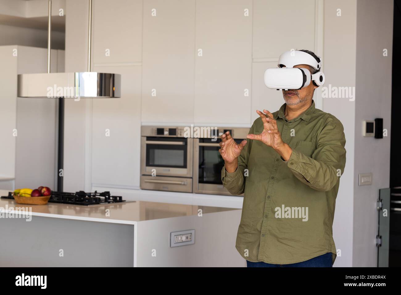 Reifer birassischer Mann mit VR-Headset, Interaktion mit virtuellen Objekten, zu Hause, Kopierraum. Moderne Küche mit eleganten Geräten und einer Schüssel Obst Stockfoto