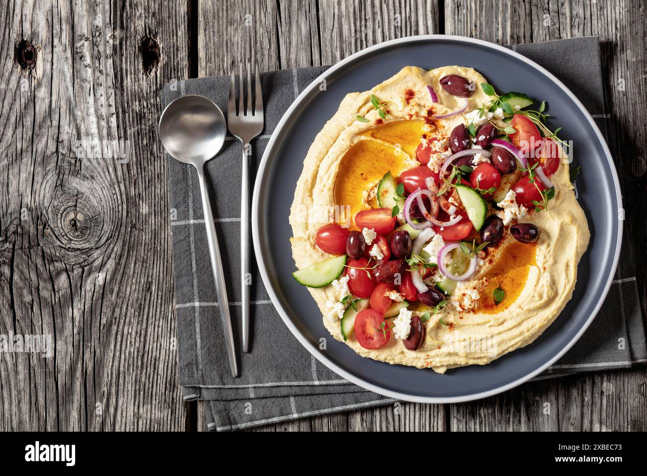 Griechischer Hummus mit frischen Gurken, Oliven, Tomaten, Fetakäse, roten Zwiebeln und Thymian auf Teller auf rustikalem Holztisch mit Löffel und Gabel, Hor Stockfoto