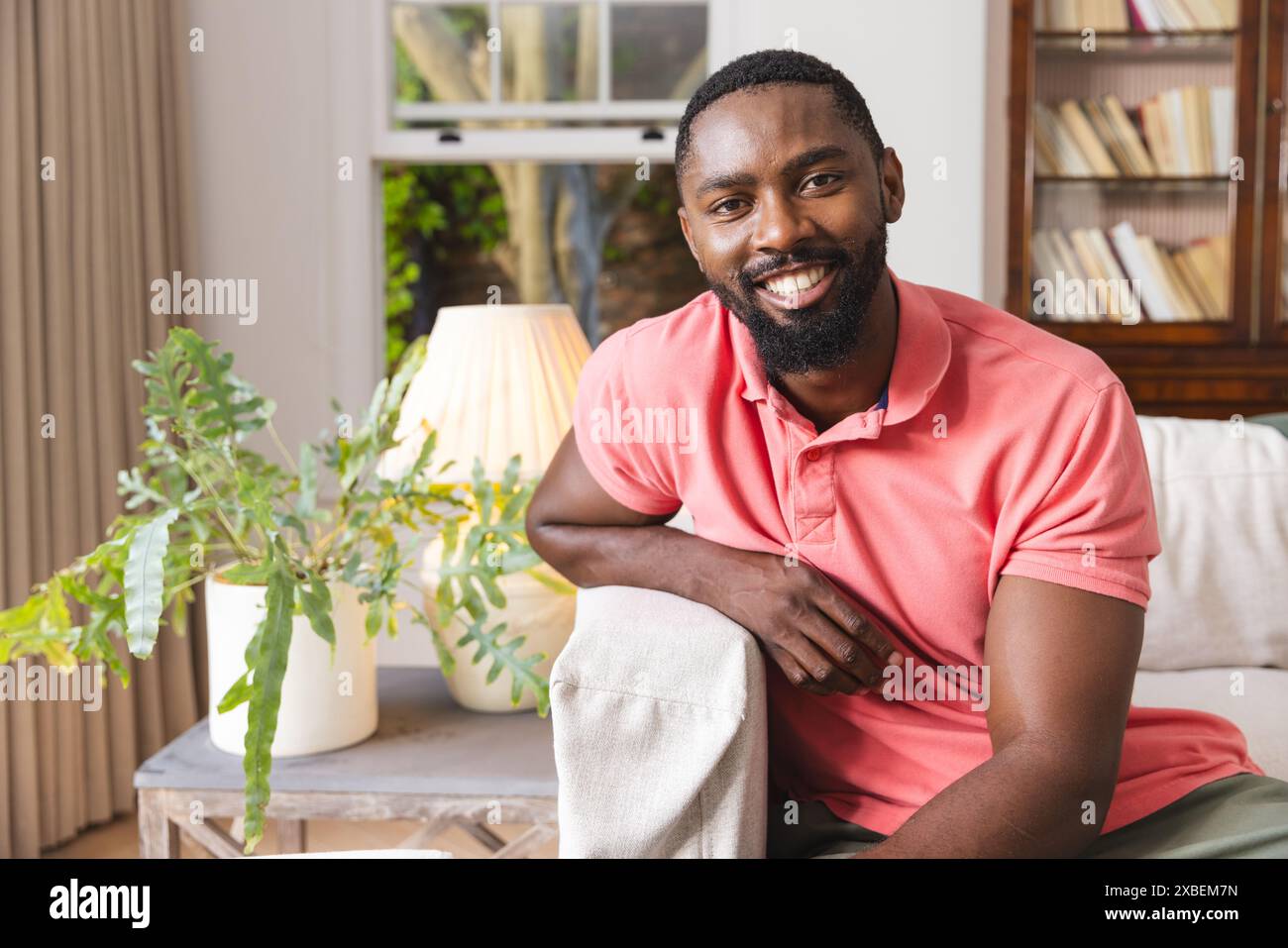 Junger afroamerikanischer Mann lächelt, während er auf der Couch im gemütlichen Wohnzimmer sitzt, zu Hause Stockfoto