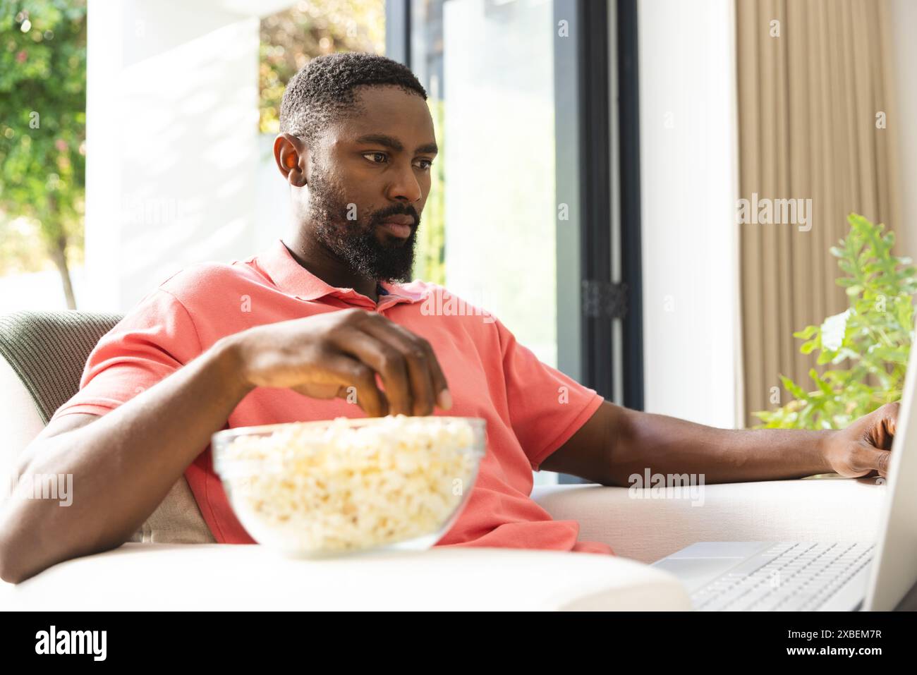 Junger afroamerikanischer Mann, der sich zu Hause entspannt, Popcorn isst, während er Laptop benutzt, zu Hause. Helles, modernes Wohnzimmer mit großen Fenstern und Grünflächen Stockfoto