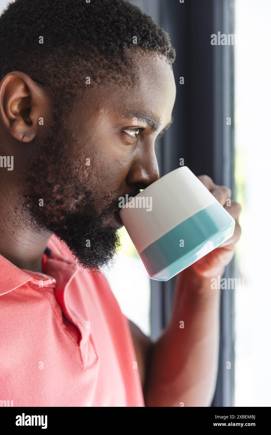 Junger Mann aus Afroamerikaner trinkt aus weißem und blauem Becher am Fenster, zu Hause Stockfoto