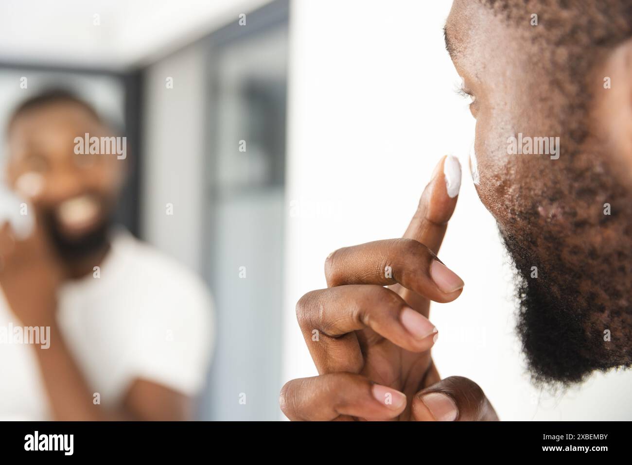Junger afroamerikanischer Mann, der Hautpflegecreme aufträgt und sein Spiegelbild anlächelt, zu Hause. Helles, modernes Badezimmer mit Tageslicht in die Stockfoto