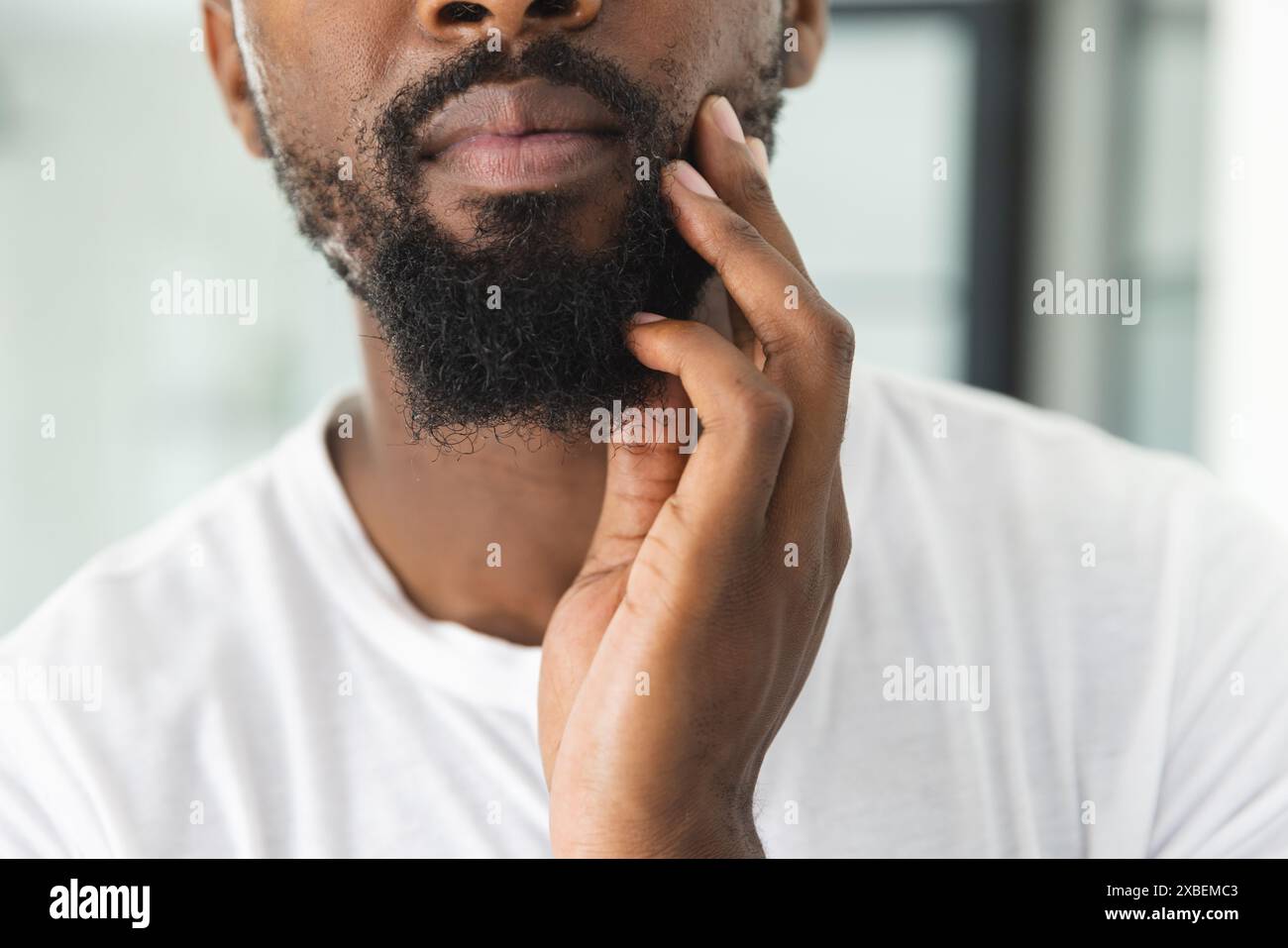 Junger afroamerikanischer Mann, der nachdenklich seinen Bart berührt, zu Hause Stockfoto