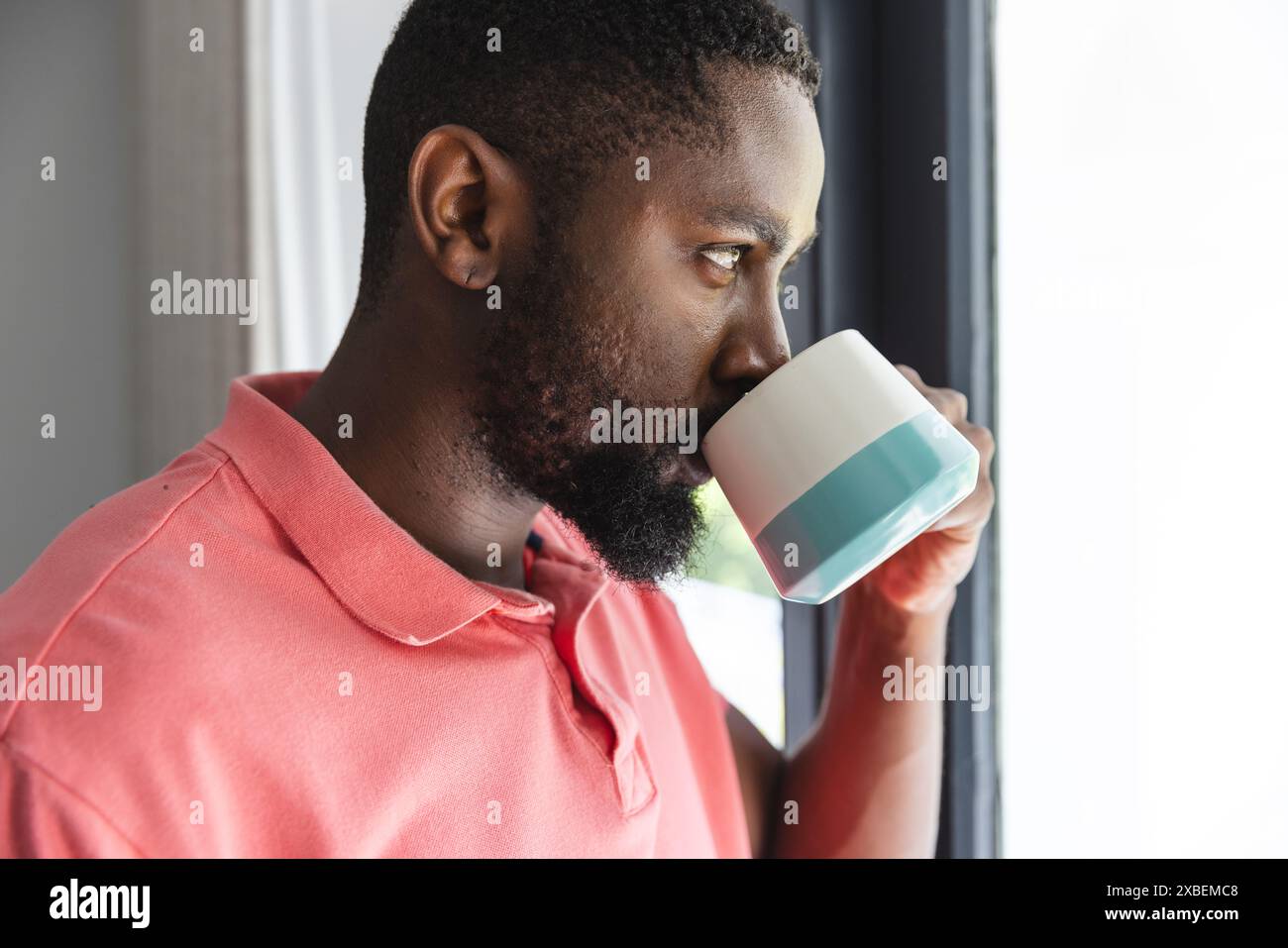 Ein junger Mann aus Afroamerikaner trinkt Kaffee, während er aus dem Fenster blickt, zu Hause Stockfoto