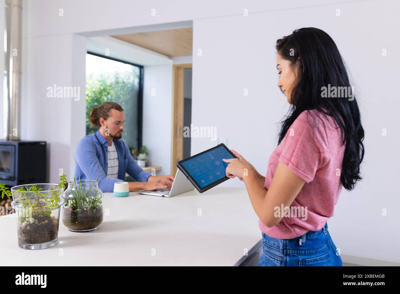 Junge birassische Frau, die ein Tablet mit Smart Home benutzt, kaukasischer Mann, der am Laptop arbeitet. Moderne Küche mit großen Fenstern und Topfpflanzen auf der Theke, un Stockfoto