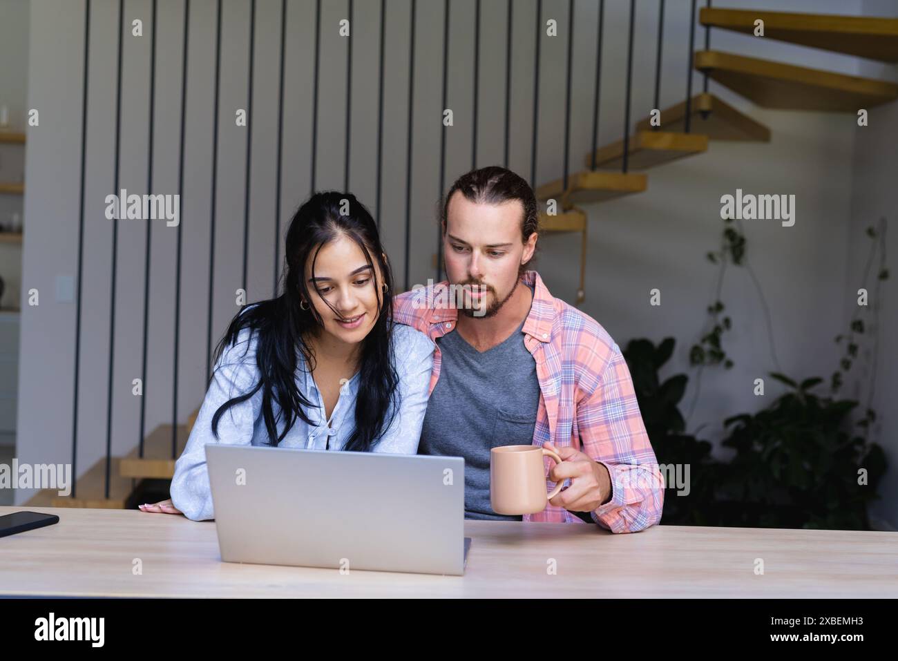 Ein Paar sitzt zusammen und sieht auf einen Laptop-Bildschirm, den Mann mit einem Becher. Sie sind in einem modernen Haus mit einer Holztreppe und Pflanzen im Hinterland Stockfoto