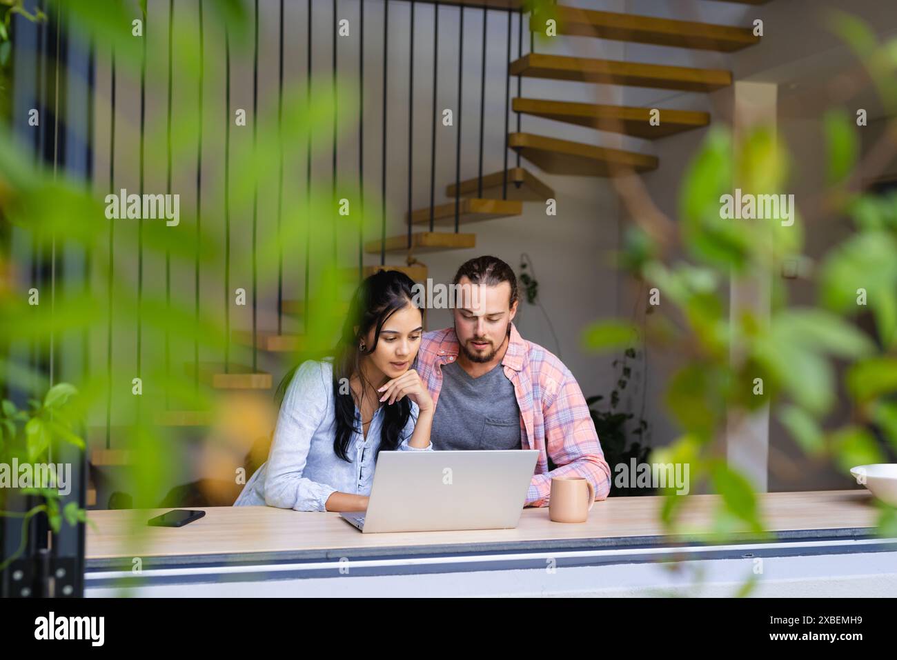 Ein vielseitiges Paar, das zusammen sitzt und auf den Laptop-Bildschirm blickt. In einem modernen Haus mit einer stilvollen Treppe und Grün, die draußen sichtbar ist, schaffen eine ruhige Atmosphäre Stockfoto