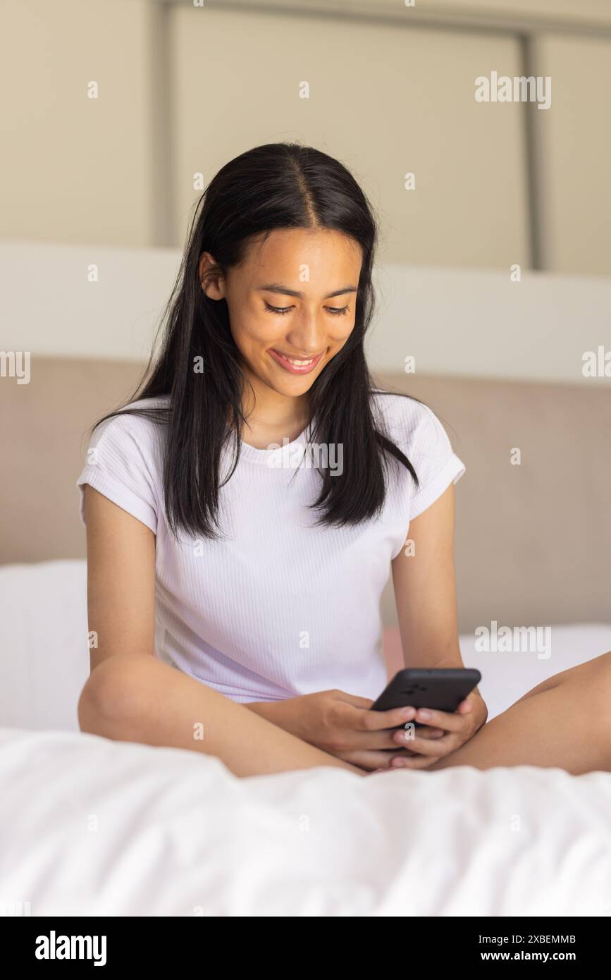 Birassische junge Frau, die auf dem Bett sitzt, ihr Smartphone lächelt, zu Hause Stockfoto