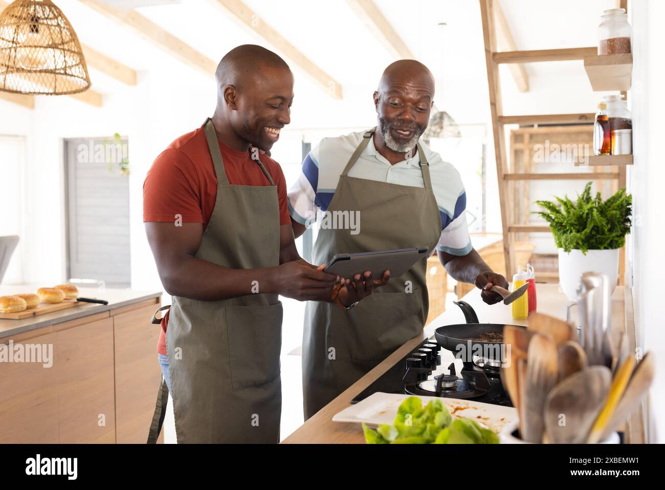 Afroamerikanischer Vater und Sohn kochen zusammen, lächeln und sehen sich die Tafel an. Moderne Küche mit Holzakzenten und frischen Zutaten auf der Theke, Stockfoto