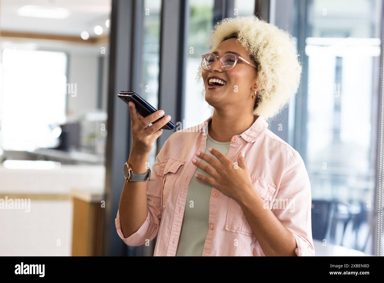 Junge birassische Frau, die Smartphone hält und im Büro lächelt. Glastrennwände und moderne Einrichtung schaffen einen eleganten, unveränderten Hintergrund Stockfoto