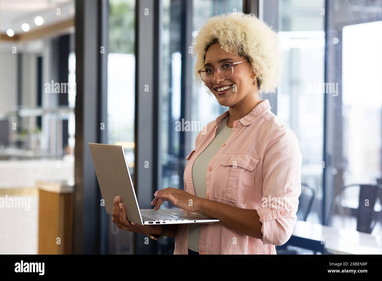 Birassische junge Frau, die einen Laptop hält und selbstbewusst in der Büroumgebung lächelt. Moderner Arbeitsbereich mit Glasabtrennungen und Tageslicht, Stockfoto