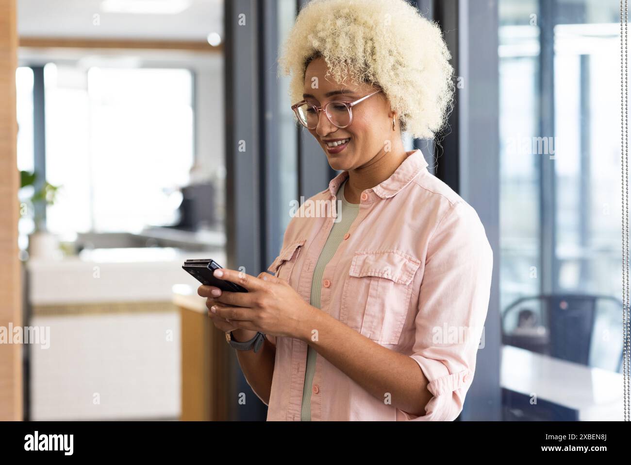 Junge birassische Frau mit lockigem Haar mit Smartphone im Büro. Moderner Arbeitsbereich mit Glaswänden und Tageslicht, der helle und helle Räume schafft Stockfoto