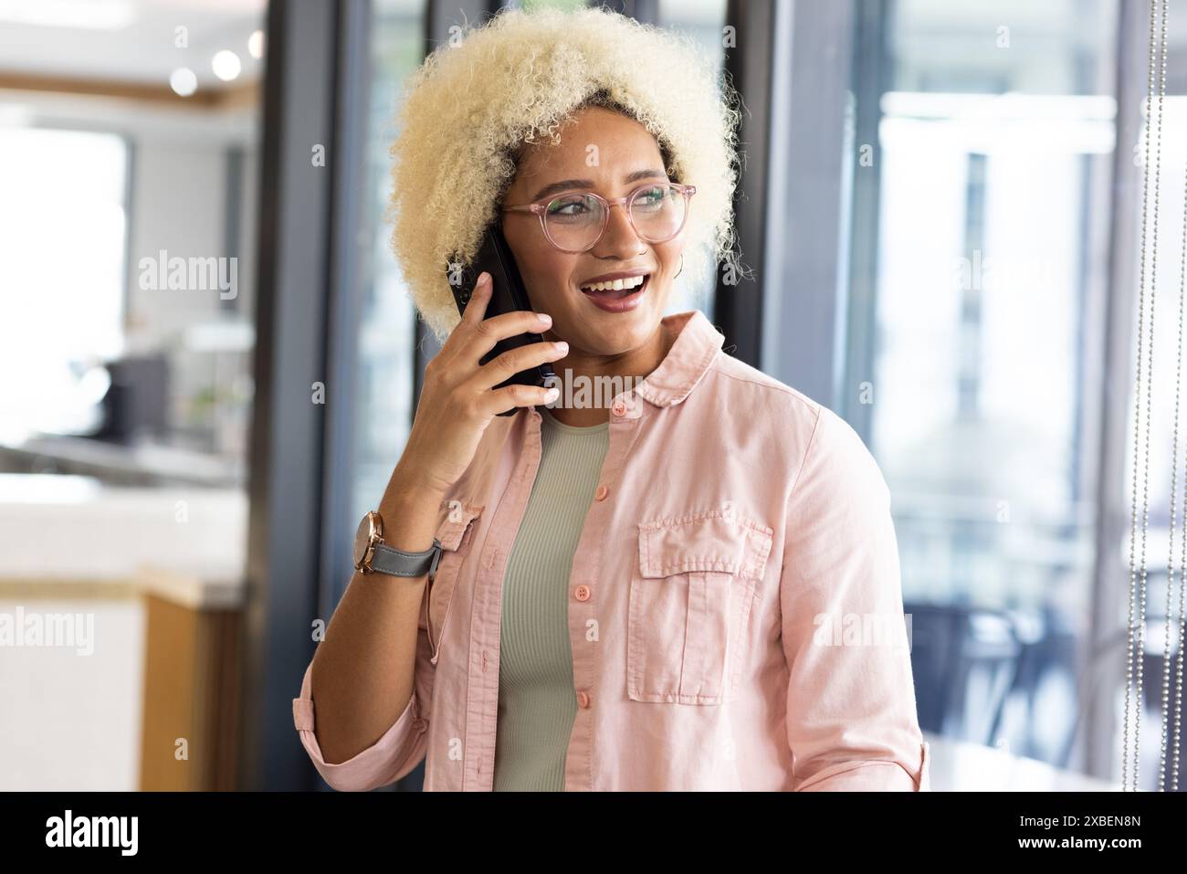 Birassische Frau mit lockigem Haar, Brille, in Freizeitkleidung, mit Smartphone. Mit Glaswänden und modernen Möbeln im Hintergrund, unverändert Stockfoto