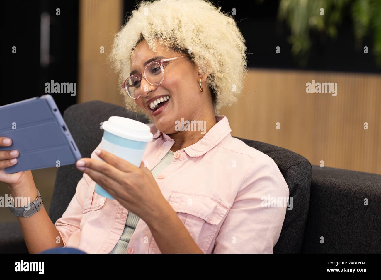Junge Frau im Büro, die Tablette und Kaffeetasse hält, lächelnd. Sitzgelegenheit auf Couch im modernen Business-Office-Loungebereich mit Holzvertäfelung und Stockfoto