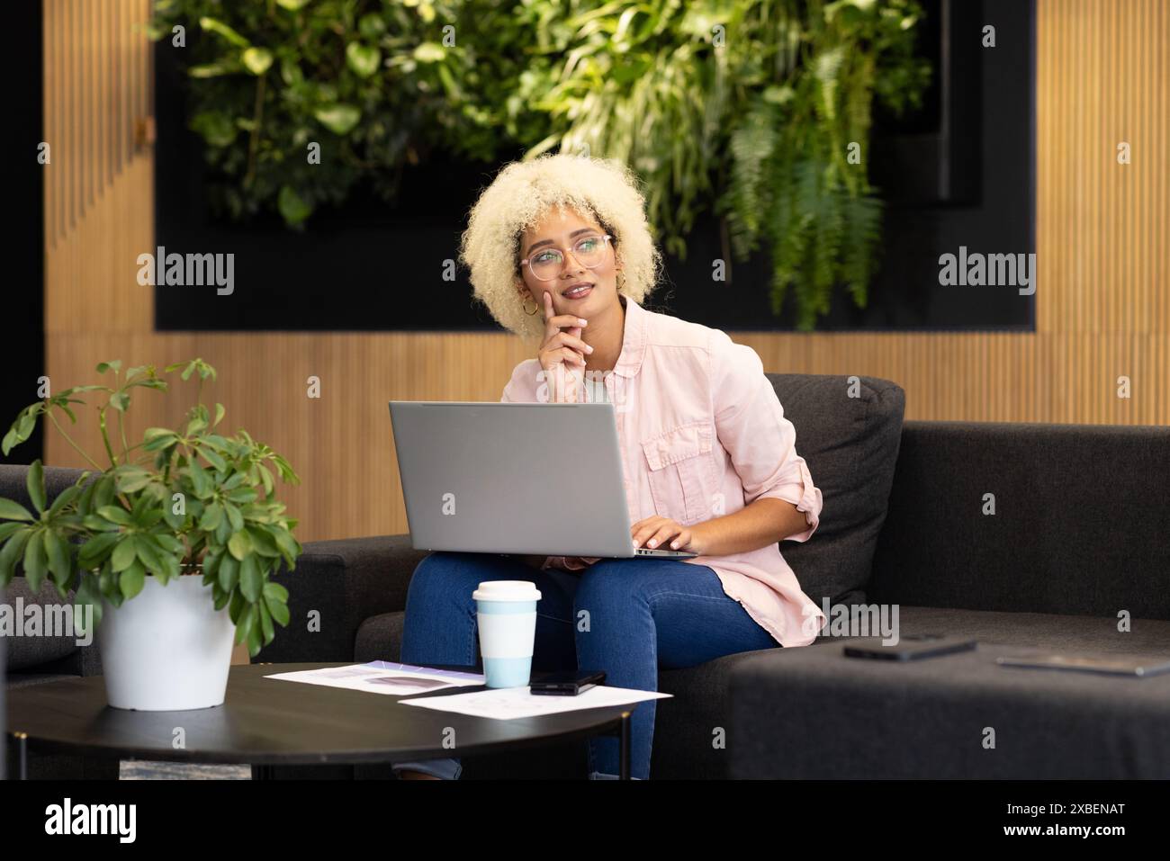 Birassische junge Frau im Büro mit Laptop, während sie auf der Couch sitzt. Arbeiten an einem Projekt in einem modernen Geschäftsbüro mit Pflanzen und Holzakzenten, u Stockfoto