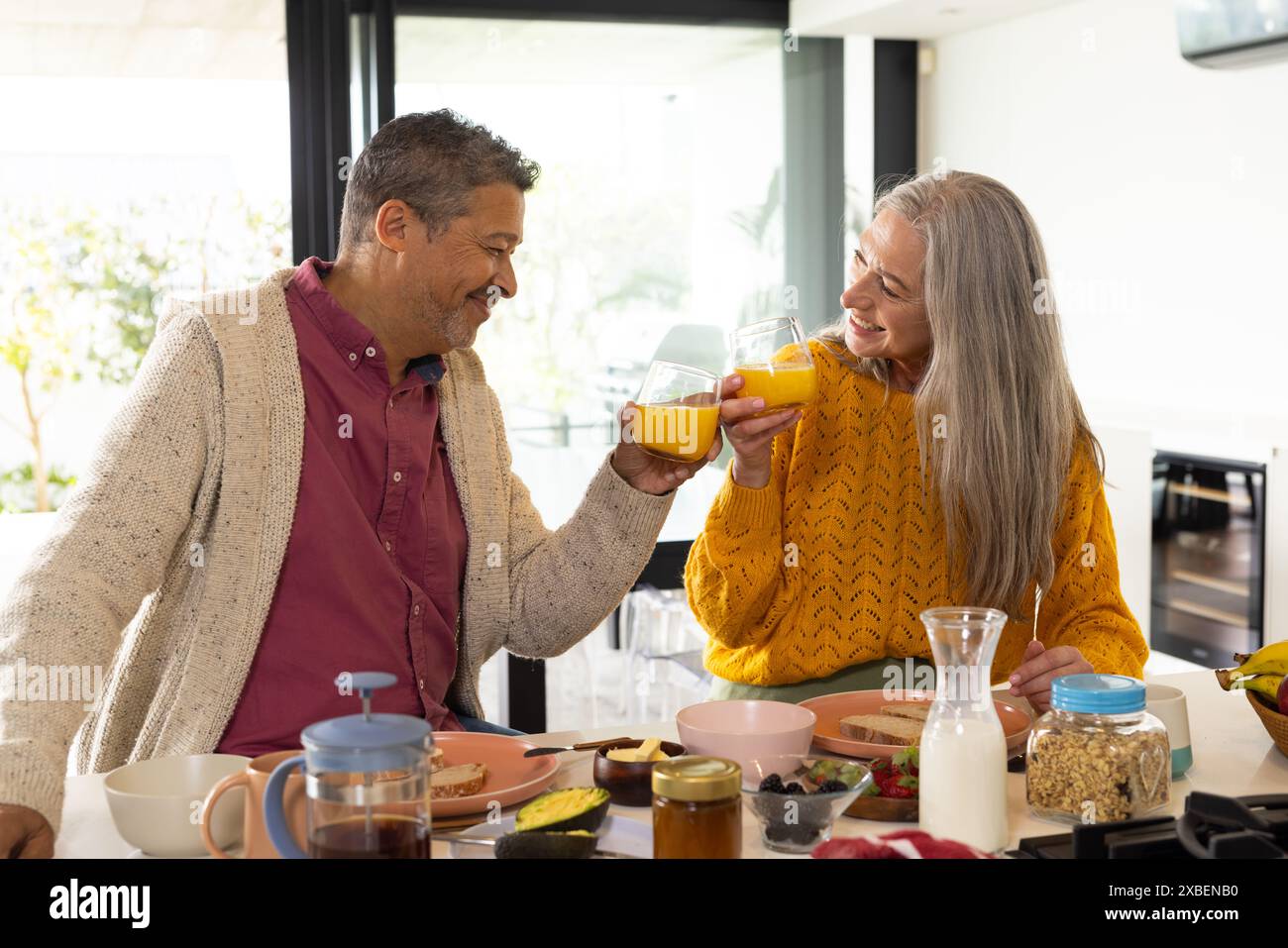 Ein zweirassiges reifes Paar lächelt und toast mit Saft beim Frühstück zu Hause, zu Hause. Helle Küche mit modernen Geräten und einer Auswahl an Frühstück Stockfoto
