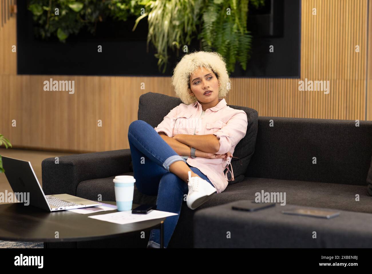 Junge birassische Frau auf Couch mit Laptop und Kaffeetasse im modernen Business Office. Holzwände und Grün im Hintergrund schaffen eine ruhige Atmosphäre Stockfoto