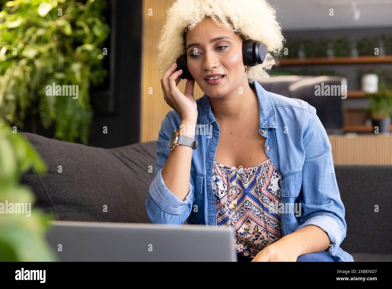 Junge birassische Frau mit Kopfhörern, die im modernen Büro auf den Laptop-Bildschirm schaut. Pflanzen und Holzakzente schaffen einen ruhigen und natürlichen Hintergrund Stockfoto