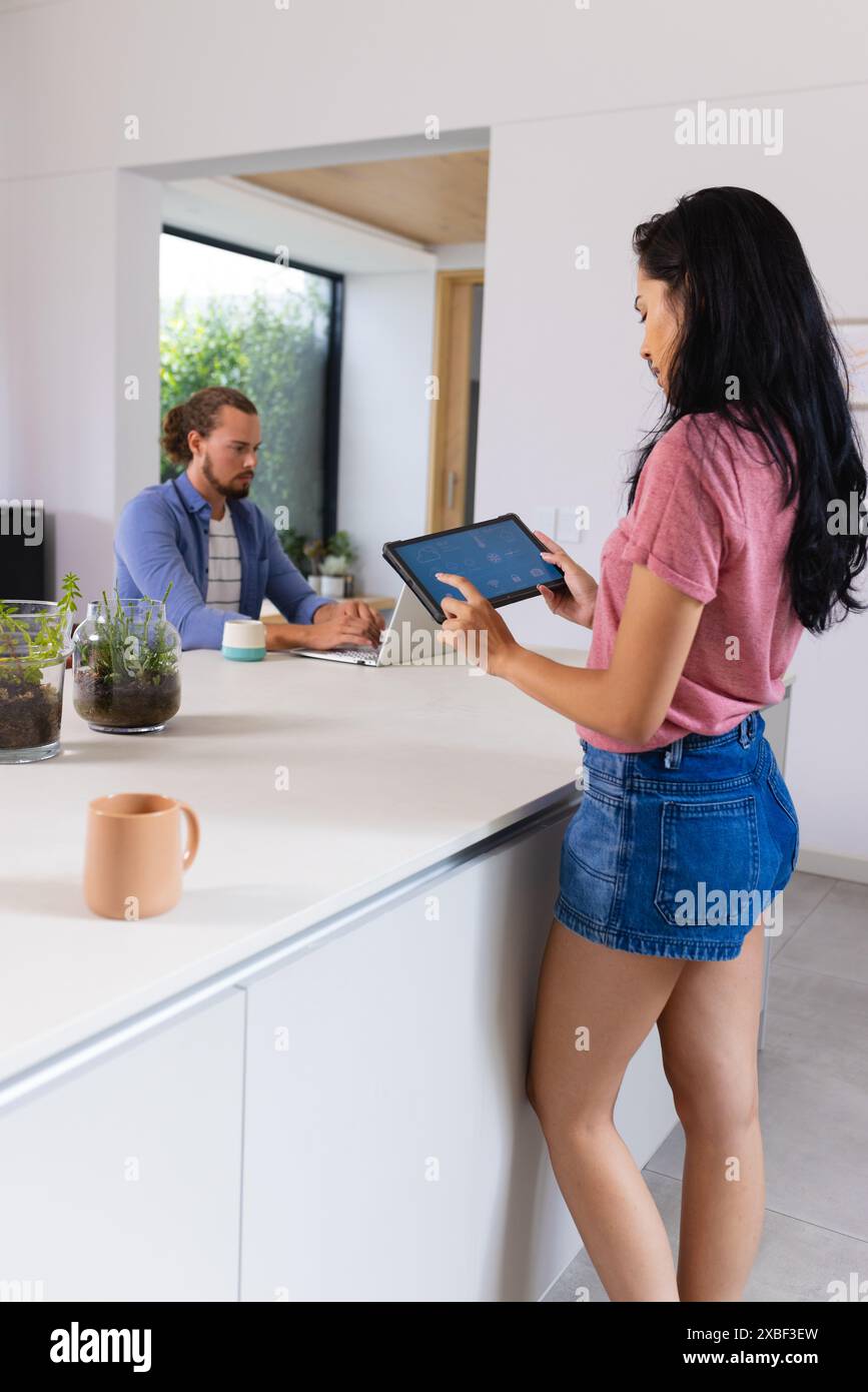 Junge birassische Frau, die ein Tablet mit Smart Home App benutzt, kaukasischer Mann, der an einem Laptop arbeitet. Helle Küche mit Pflanzen und großen Fenstern, die luftig sind Stockfoto