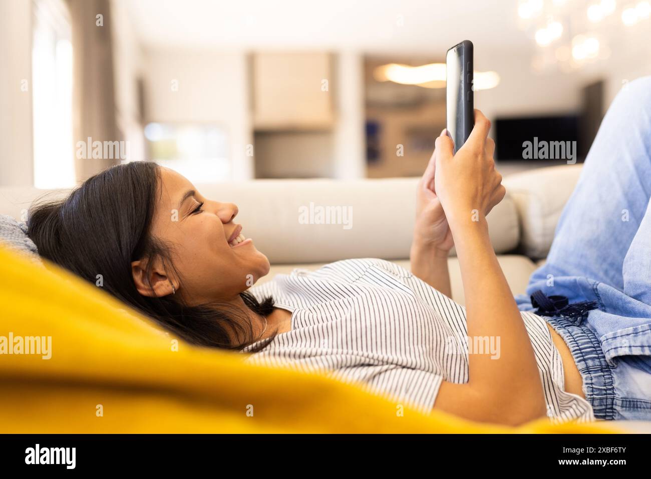 Birassische junge Frau, die sich auf der Couch entspannt, lächelt, während sie ihr Smartphone ansieht, zu Hause Stockfoto