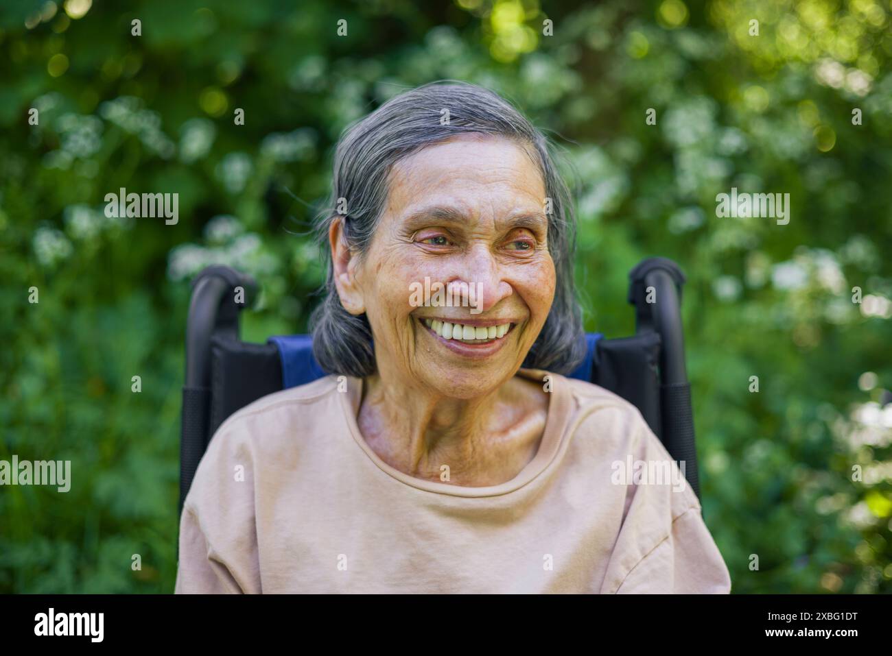 Alte indische (südasiatische) Frau in ihren 90ern lächelnd, in lässiger Kleidung sitzend in einem Rollstuhl draußen in der Natur, Großbritannien Stockfoto