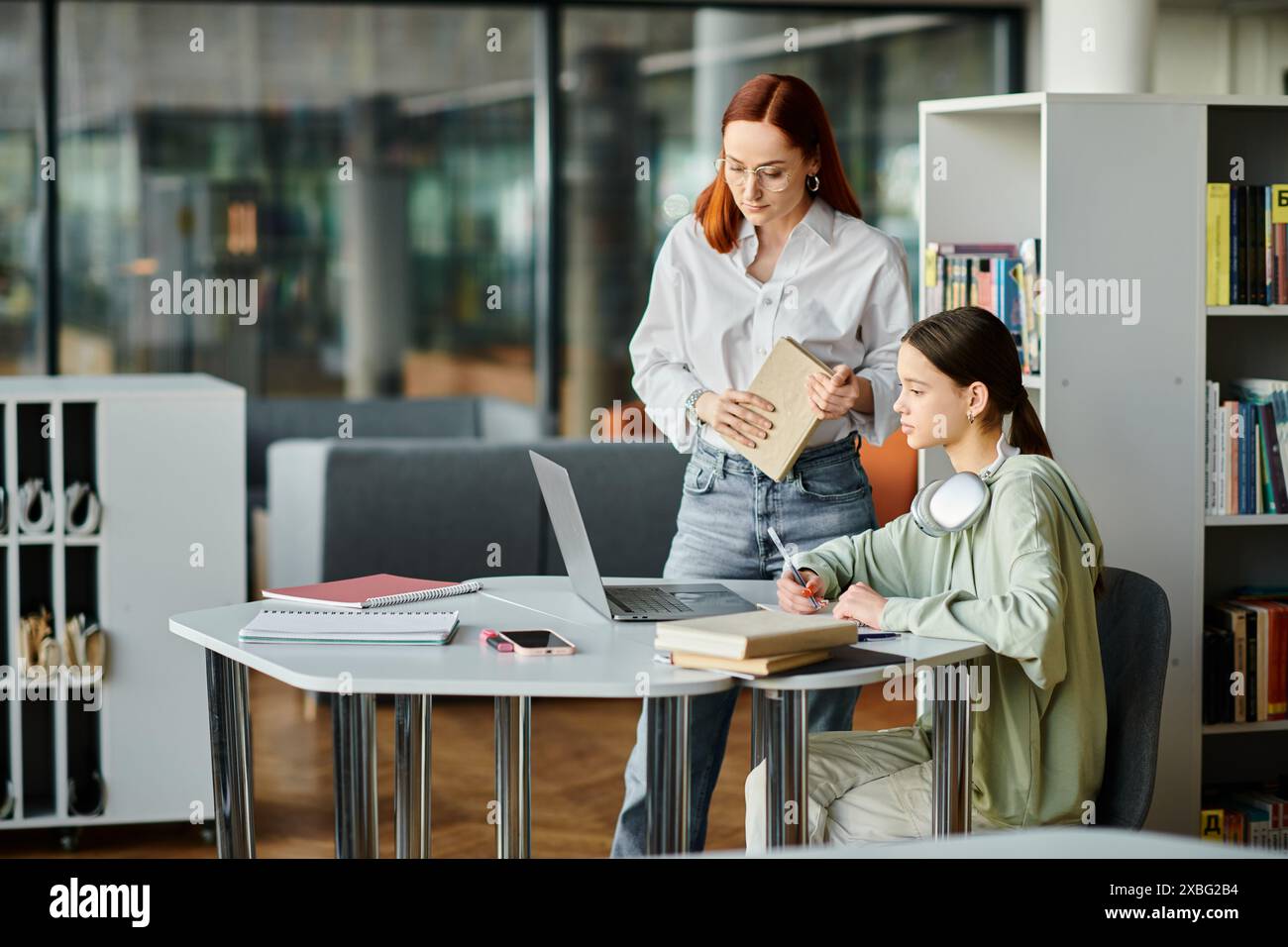 Eine rothaarige Frau unterrichtet ein Mädchen im Teenageralter in einer Bibliothek und arbeitet an einem Laptop für den Unterricht nach der Schule zusammen. Stockfoto