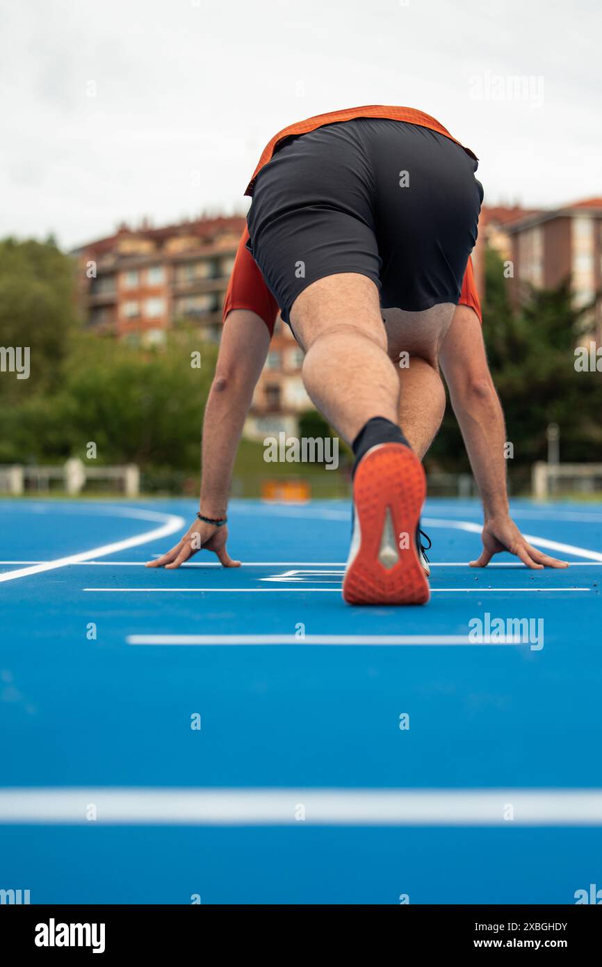 Der vertikale Läufer wird von hinten an der Startlinie gehockt und bereit für den Start auf einer blauen Leichtathletikstrecke gesehen. Dieses Bild wird aufgenommen Stockfoto