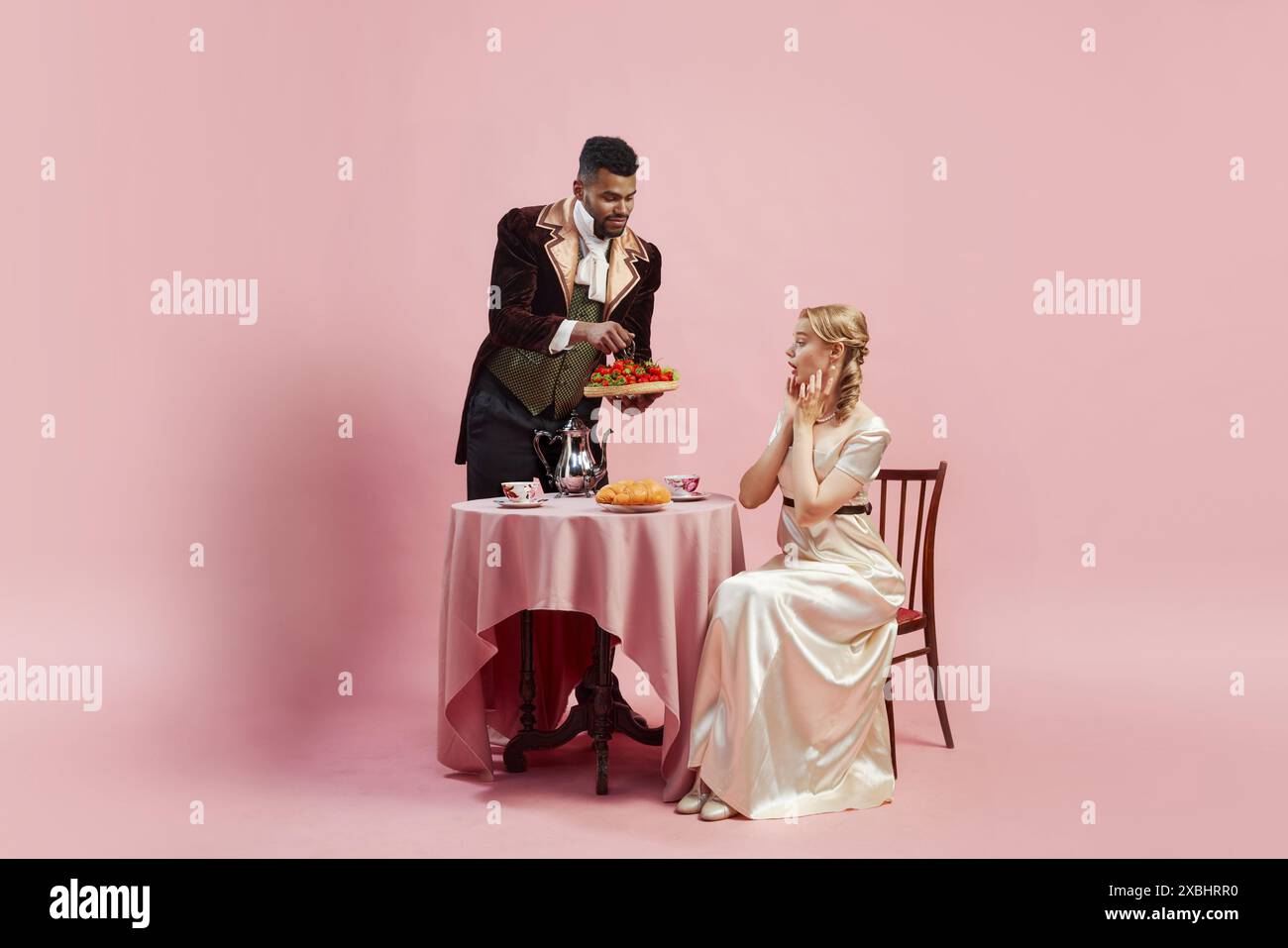 Mann in reicher Samtjacke bietet Tablett mit frischen Erdbeeren für elegante Frau in Satinkleid vor rosa Studiohintergrund Stockfoto