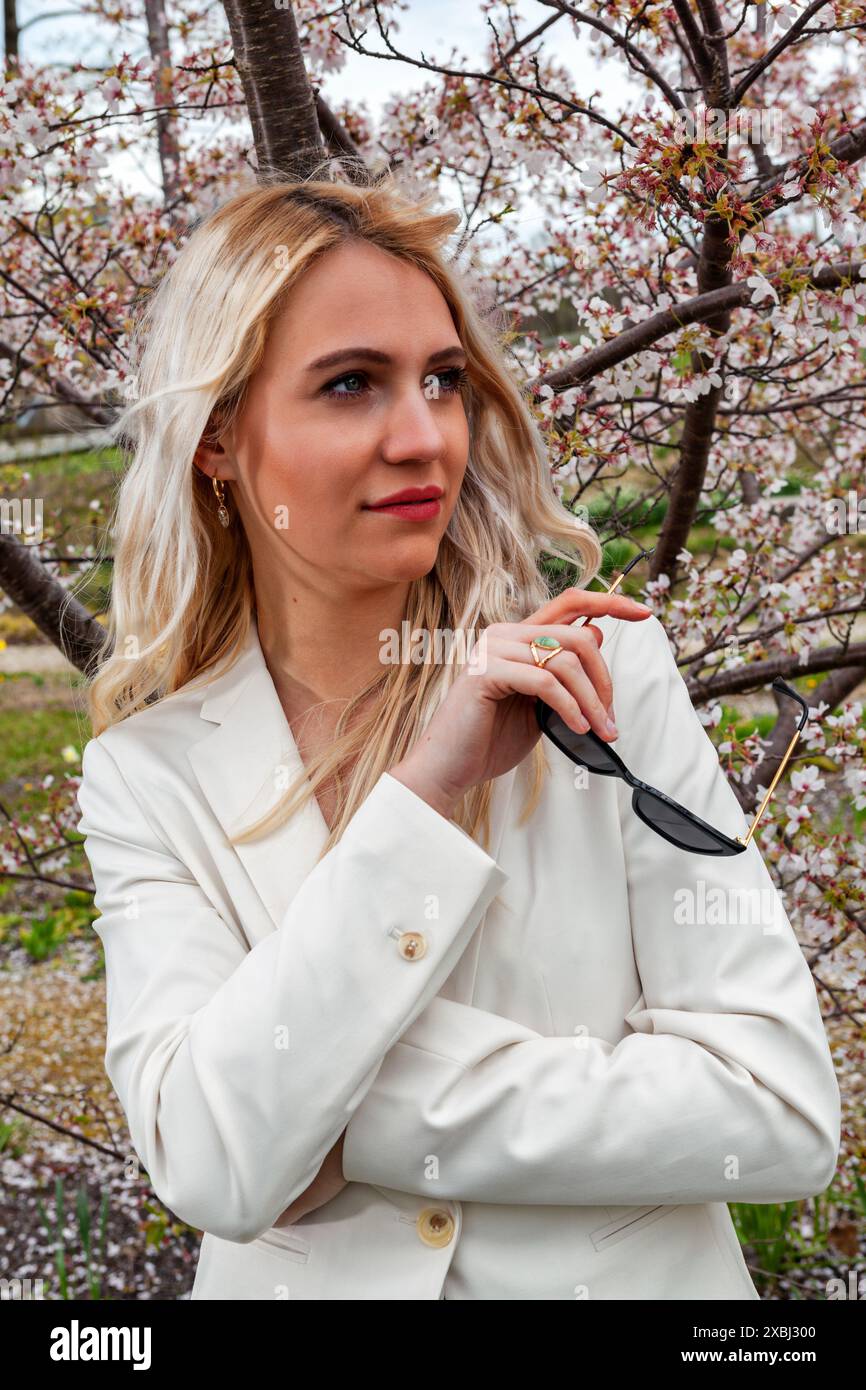 Ein Porträt einer jungen, blonden Frau, die eine Sonnenbrille hält. Im Hintergrund ist ein blühender Sträucher, der eine frühlingshafte und lebhafte Stimmung erzeugt. Die Stockfoto