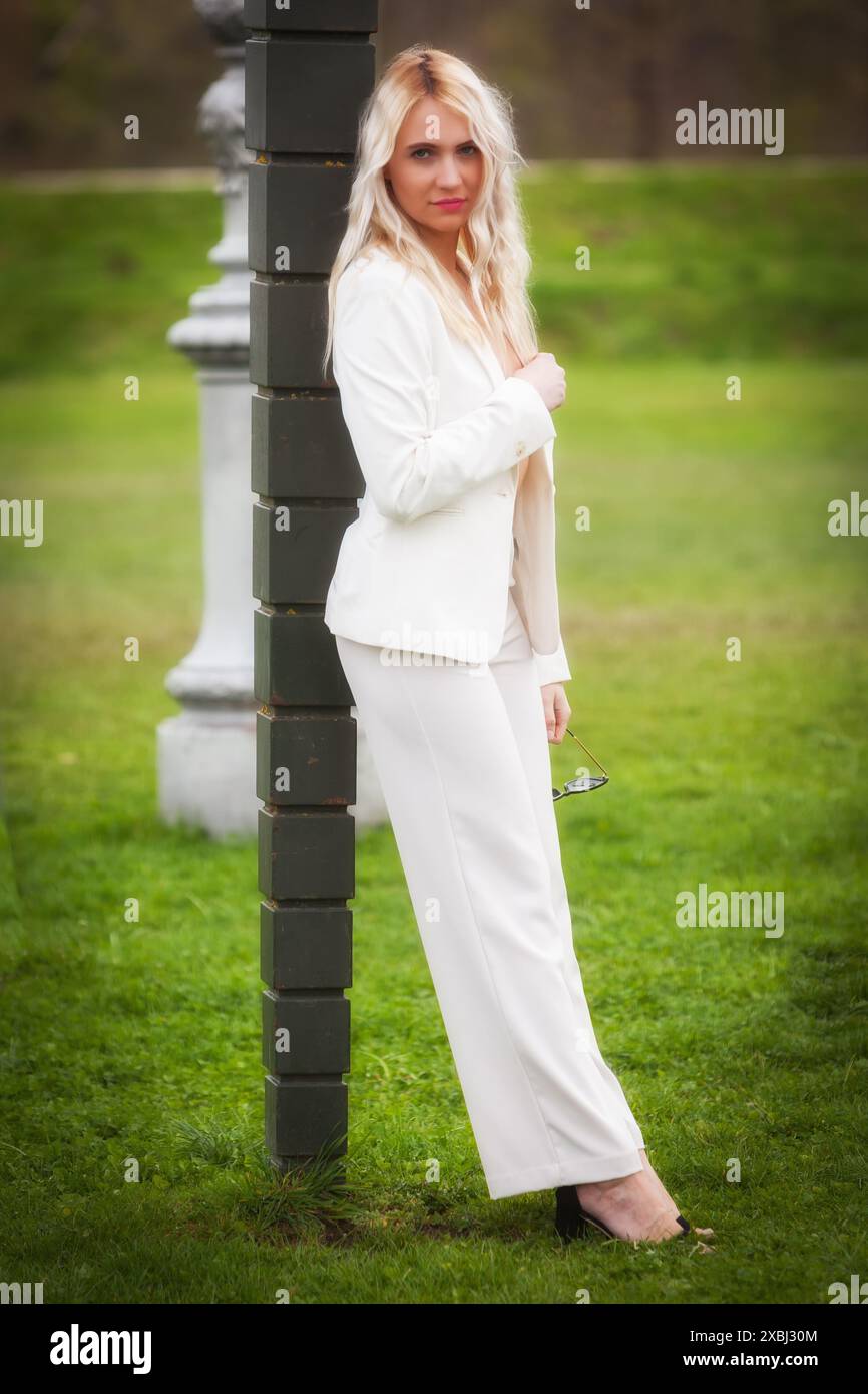 Eine junge, blonde Frau in einem weißen Hosenanzug lehnt sich an einen Laternenpfahl mitten auf einer Wiese und schaut direkt in die Kamera. Ihre selbstbewusste Position Stockfoto