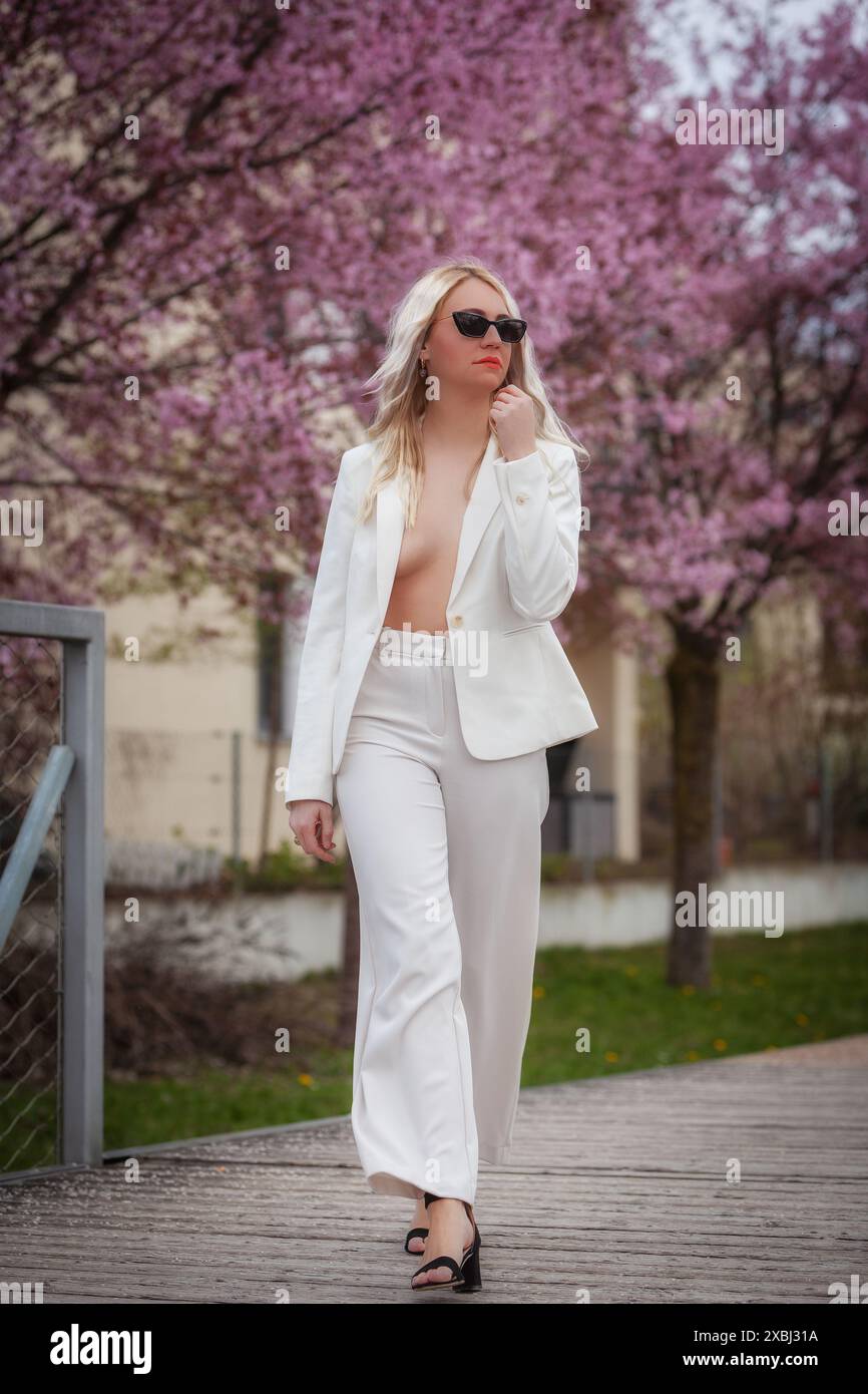 Ein Model trägt einen weißen Hosenanzug mit einer offenen Jacke, die ihre Brust zeigt, und spaziert durch einen Park voller blühender Bäume. Sie trägt eine Sonnenbrille Stockfoto