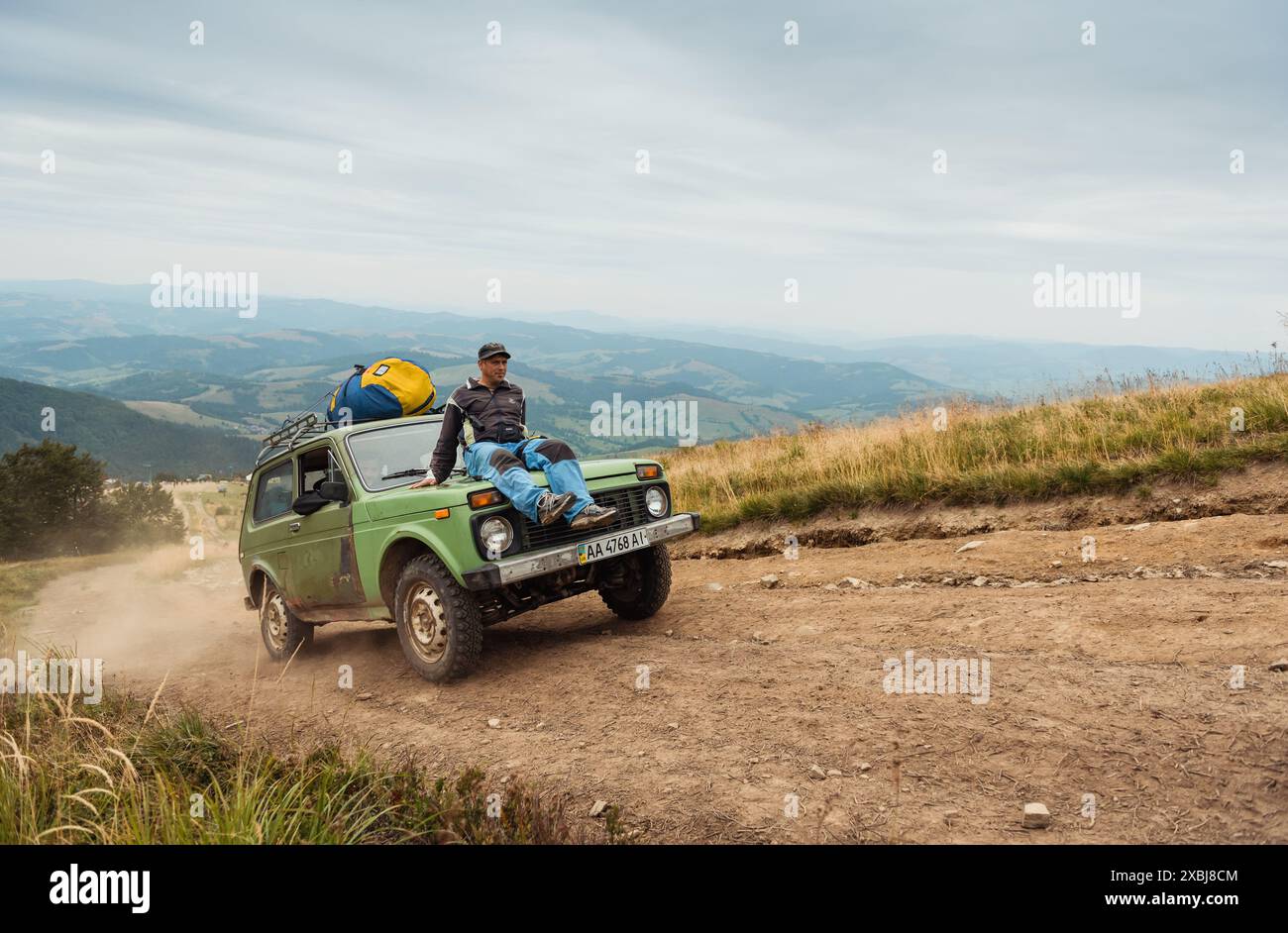 Lustige und gefährliche Fahrt in den Bergen. Ein älterer Mann sitzt auf der Motorhaube des grünen alten SUV, der bergauf fährt und Staub aufwirbelt. Stockfoto