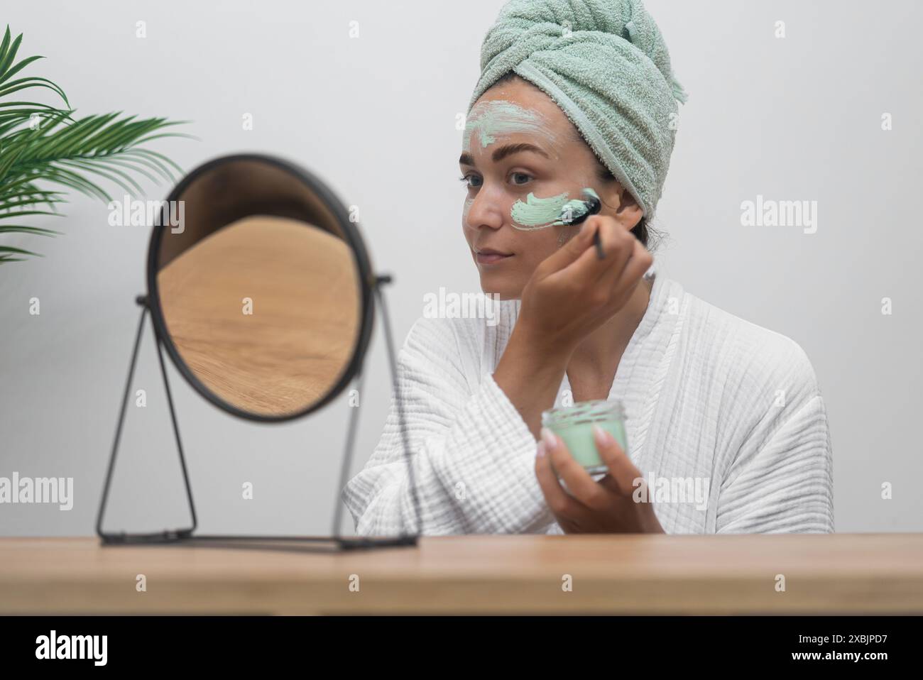 Spa at Home: In einem Bademantel trägt sie mit einer Bürste eine Tonmaske auf und blickt in den Spiegel Stockfoto