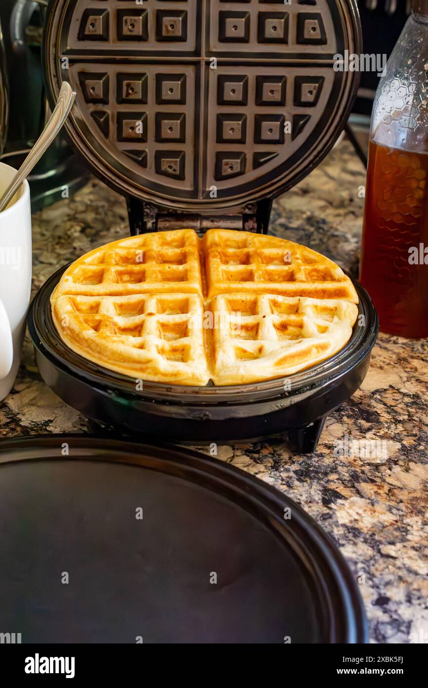 Auf dem Waffeleisen befindet sich eine fertige Waffel mit Teigbecher und Honig im Hintergrund. Stockfoto