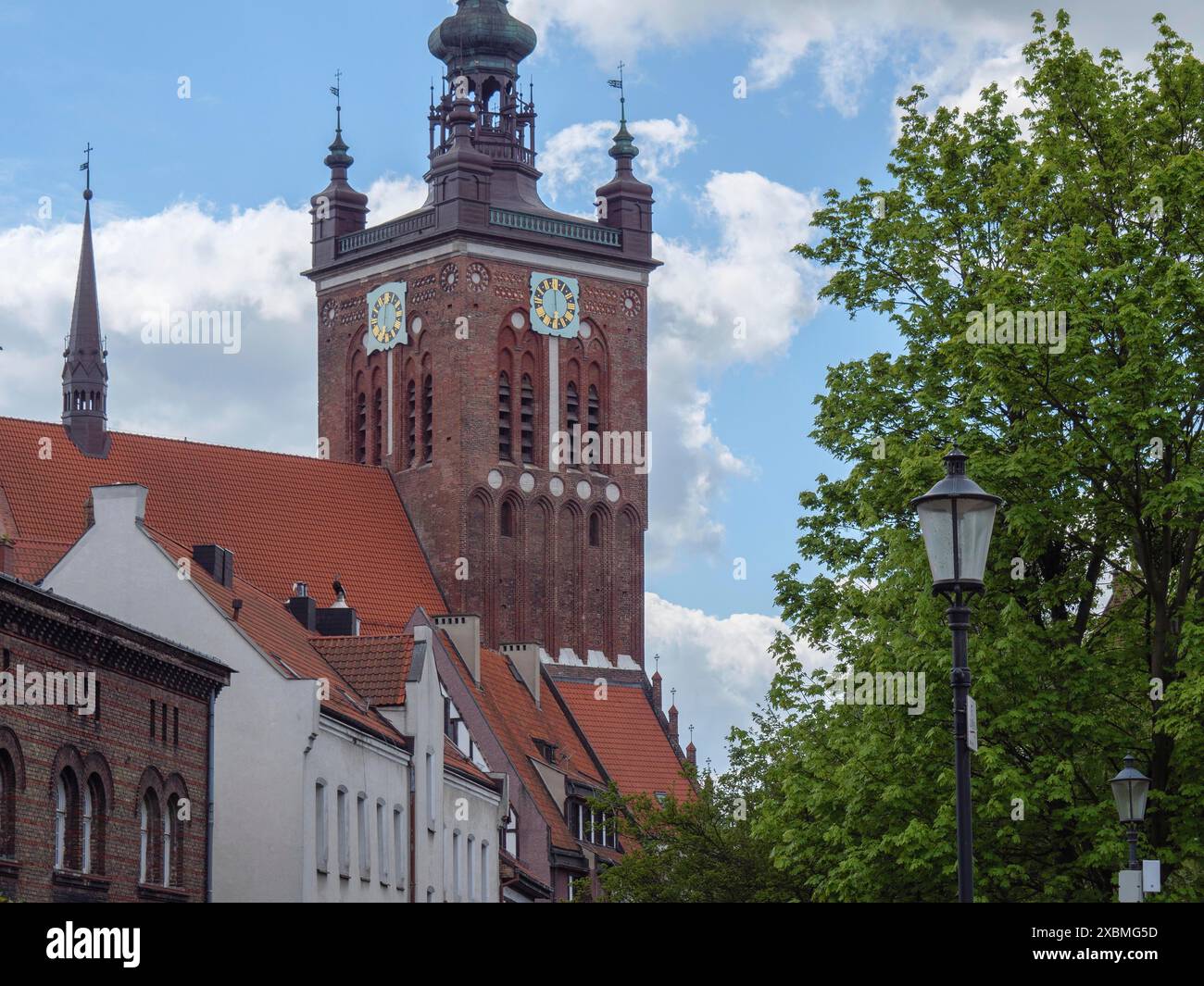 Ein gotischer Kirchturm mit roten Ziegeldächern, umgeben von grünen Bäumen und einem Lampenpfosten, Danzig, Polen Stockfoto