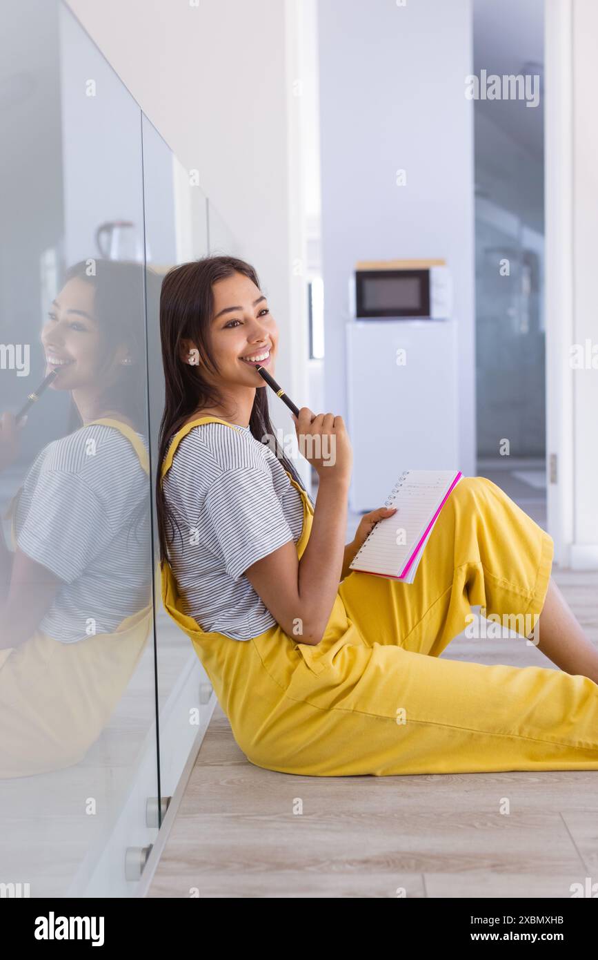 Glückliche Frau, die auf dem Boden sitzt, Notizbuch und Stift hält und nachdenklich lächelt Stockfoto