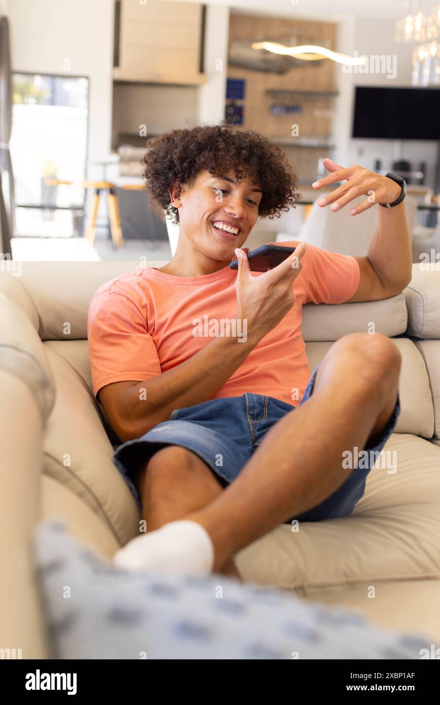 Glücklicher birassischer Mann, der sich auf der Couch entspannt und mit seinem Smartphone interagiert Stockfoto